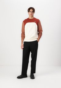 Herre fargeblokk sweatshirt i krem og rust, med ribbestrikkede mansjetter og rund hals. Kombinert med svarte bukser og svarte sko.