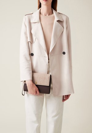 Giacca in suede beige chiaro con colletto a risvolto, due bottoni neri; tiene in mano una borsa a tracolla bicolore, parte superiore testurizzata e parte inferiore marrone. Pantaloni bianchi.