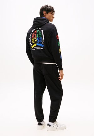 Felpa nera con logo multicolore sul retro; pantaloni della tuta neri; il modello indossa sneakers bianche con accentuazioni nere. Abbigliamento casual e sportivo.