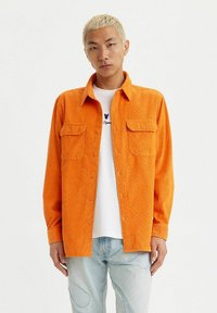 Camicia in corduroy arancione con colletto a punta, dotata di due tasche sul petto e taglio dritto, indossata sopra una maglietta bianca e jeans chiari.