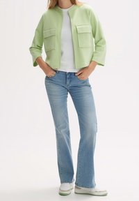 Veste courte vert clair avec deux poches avant, associée à un jean flare bleu et des baskets blanches avec des accents verts.
