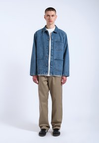 Giacca di jeans di colore azzurro chiaro con chiusura a bottoni, due tasche sul petto e una vestibilità comoda, abbinata a pantaloni beige e sneakers nere.