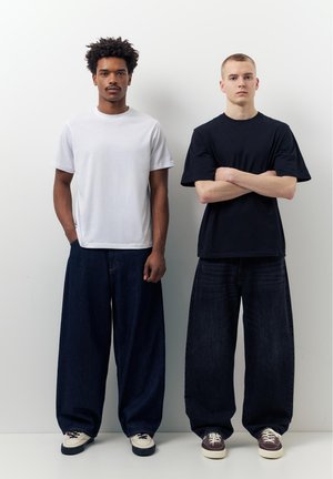 Doi bărbați stau unul lângă altul, purtând tricouri simple albe și negre, blugi largi întunecați și adidași casual, pe un fundal alb simplu.