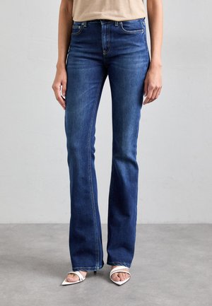 Jeans di denim blu scuro a zampa d'elefante con una texture liscia, caratterizzati da dettagli di cucitura sottile e vita media. Abbinati a sandali bianchi con tacco.
