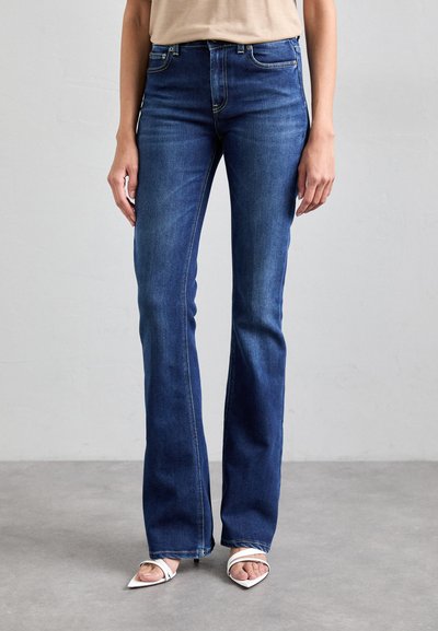 Jeans en denim bleu foncé évasés avec une texture lisse, présentant des détails de couture subtils et une taille mi-haute. Associés à des sandales à talons blancs.