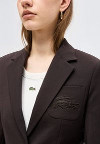Femme portant un blazer noir avec un logo de crocodile sur la poche et un haut blanc avec un petit emblème de crocodile vert, une boucle d'oreille dorée visible.