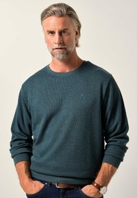 Maglione a maniche lunghe color teal scuro con texture a coste e scollo rotondo. Presenta una piccola lettera "B" ricamata sul petto. Indossato con jeans scuri.
