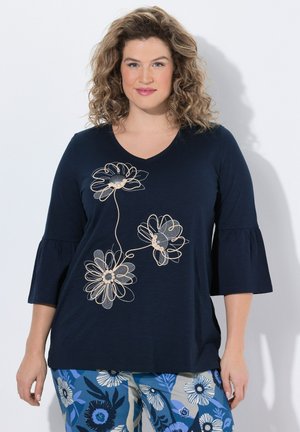 Frau mit lockigem Haar, die ein marineblaues Oberteil mit beiger Blumenstickerei und blau gemusterte Blumenhose trägt, steht vor einem weißen Hintergrund.