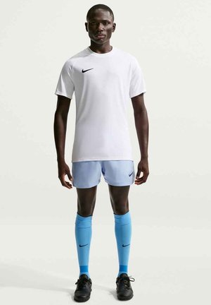 Atleta in piedi, indossa una maglietta bianca Nike, pantaloncini Nike azzurri, calzettoni blu fino al ginocchio e scarpe da calcio nere, su uno sfondo chiaro uniforme.