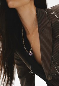 Collar de cadena de clip de papel dorado con un colgante circular púrpura, llevado sobre una blusa oscura y ajustada y un blazer a rayas. Detalles de tejido texturizado visibles.