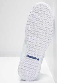 Baskets blanches avec semelle en caoutchouc texturée, présentant un motif de bande de roulement et un petit logo bleu Reebok sur le côté. Matériau supérieur lisse.