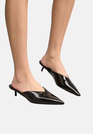Pieds portant des mules en cuir noir à bout pointu avec de petits talons kitten, présentés sur un fond clair uni.