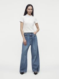 Jeune femme aux longs cheveux foncés porte un haut blanc à manches courtes et un jean bleu ample à jambes larges avec des chaussures noires pointues, debout avec une main dans la poche.