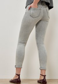 Persona vistiendo jeans skinny gris claro con los puños enrollados y zapatos planos con correa al tobillo color burdeos, con la mano en el bolsillo trasero, de pie contra una pared lisa.