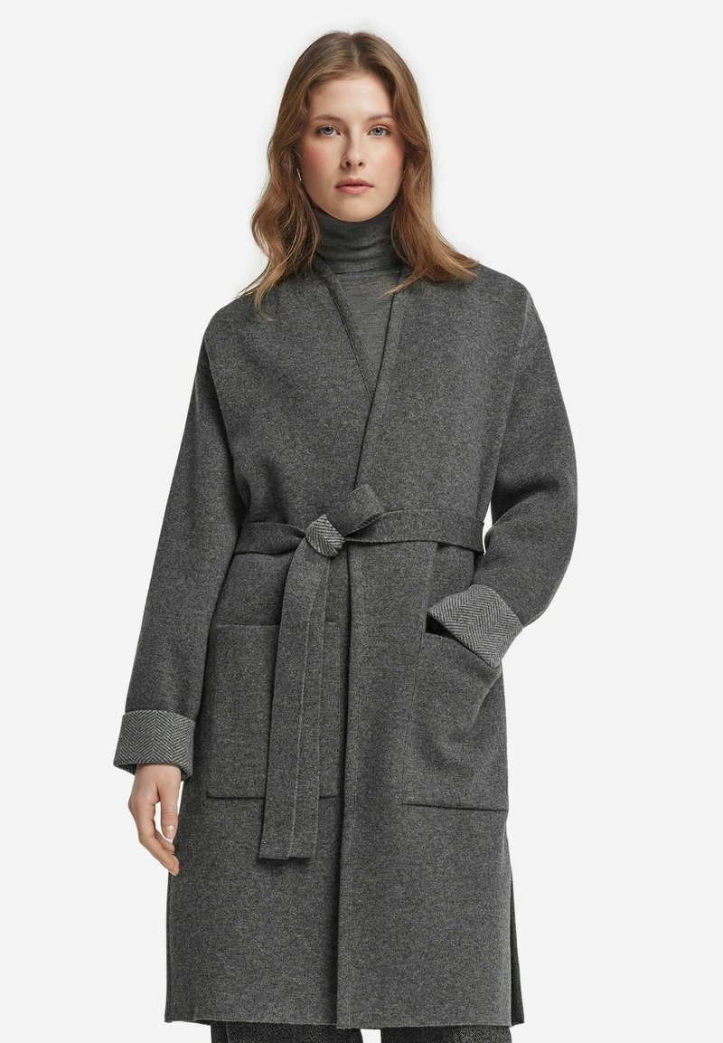 Grau melierter, langer Wollmantel mit einem Bindegürtel, der zwei Fronttaschen und Hahnentritt-musterte Bündchen aufweist. Glatte, weiche Textur.