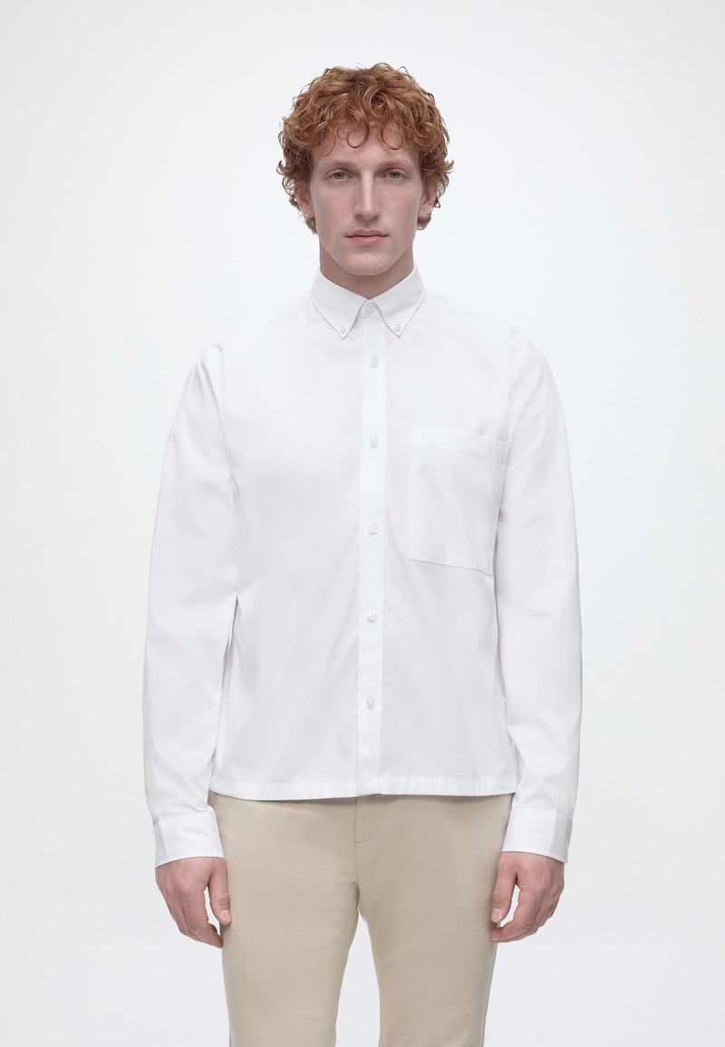 Άνδρας με σγουρά κόκκινα μαλλιά, που φοράει λευκό πουκάμισο με κουμπιά και μπεζ παντελόνι, στέκεται μπροστά σε απλό λευκό φόντο.