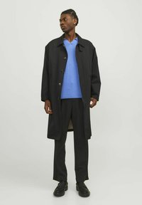 Lång svart kappa över en ljusblå V-hals tröja och mörka byxor. Modellen bär svarta skor; kännetecken inkluderar en knappfront och uttalad krage.