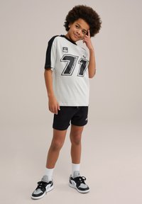 Balta t-krekls ar melniem piedurknēm, redzamā ir liela "71" grafika un teksts. Melni šorti un melni/balti kedas noslēdz apģērbu.