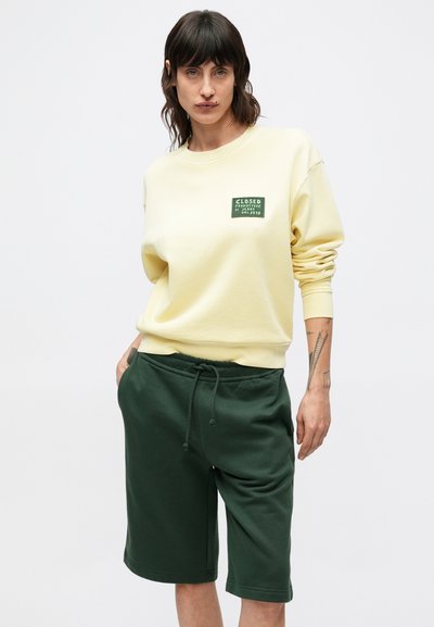 Sweat-shirt décontracté jaune avec un écusson étiquette vert, associé à un short à cordon de serrage vert foncé. Texture douce, coupe décontractée et short longueur genou.
