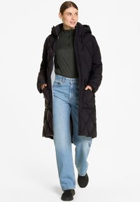 Lungo cappotto imbottito nero con cappuccio, dotato di chiusura a zip, tasche laterali e un motivo a diamante testurizzato. Indossato sopra un top scuro e jeans chiari.