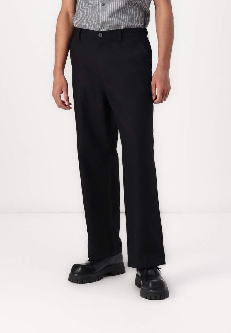 Homme portant un pantalon habillé noir et des chaussures noires à semelle épaisse, debout devant un fond blanc uni.