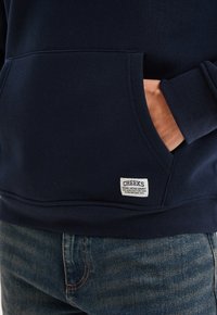 Persona vistiendo una sudadera con capucha azul oscuro, con la mano en el bolsillo delantero y una etiqueta que dice "CREEKS Original Vintage Garment" sobre unos jeans azules.