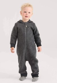 Peuter met blond krullend haar die staat, gekleed in een donkergrijze fleece jumpsuit met capuchon en grijze gebreide sokken op een lichte achtergrond.