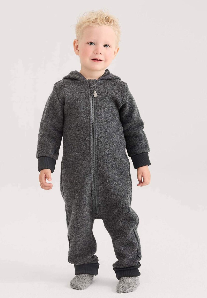 Peuter met blond krullend haar die staat, gekleed in een donkergrijze fleece jumpsuit met capuchon en grijze gebreide sokken op een lichte achtergrond.