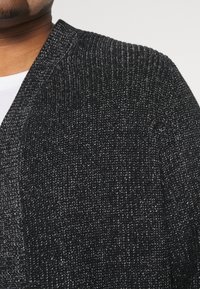 Svart texturerad kofta med en lös vävdesign, med v-ringning och synliga sömmar, som delvis täcker en vit skjorta under.