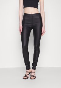 Vysoké černé legíny vyrobené z lesklého, elastického materiálu. Úzce padnoucí design s hladkým povrchem a bez viditelných vzorů. Nosič s černými páskovými podpatky.