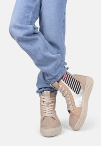 Sneakers high-top beige chiaro con dettagli in suede e accenti lucidi, caratterizzati da un dettaglio in tessuto a righe e suola in gomma beige, abbinate a jeans in denim.