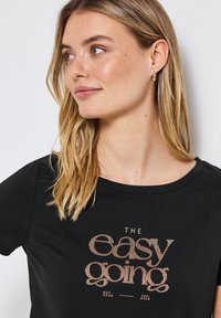 Černé tričko s kulatým výstřihem, které má grafiku s pruhovaným textem v růžovém zlatě, jež zní "THE easy going." Měkká textilie.