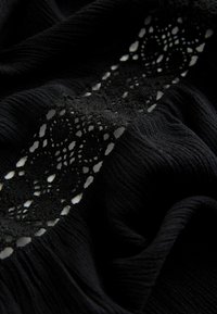 Tissu noir texturé avec une bande verticale centrale en dentelle complexe présentant des motifs en losange et en ovale, montrant des plis et des ombres.