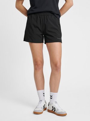 Persoon die zwarte sportshorts draagt, witte sportsokken met zwarte chevrons en witte sneakers met zwarte details, staand met de handen achter de rug.