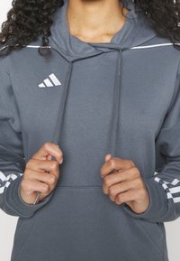 Sweat-shirt à capuche gris avec des rayures blanches sur les manches, détail de cordon de serrage et logo Adidas sur la poitrine. Tissu doux et coupe décontractée.
