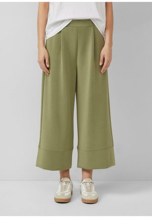 Osoba w oliwkowozielonych szerokich spodniach o kroju culotte i białych trampkach stojąca na jednolitym szarym tle.