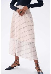 Fließender Cremefarben Rock mit schwarzem Schriftmuster, featuring einen hohen und niedrigen Saum und kombiniert mit schwarzen Absatzschuhen, die mit Perlenakzenten verziert sind.