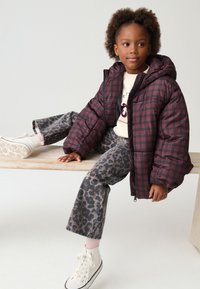 Blouson matelassé à carreaux bordeaux avec capuche, jean gris à imprimé léopard, et baskets montantes blanches. Le mannequin est assis sur une surface en bois.