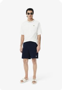 T-shirt blanc à manches courtes avec un col rond, short bleu marine et sandales crème à enfiler. Des lunettes de soleil réfléchissantes complètent la tenue.