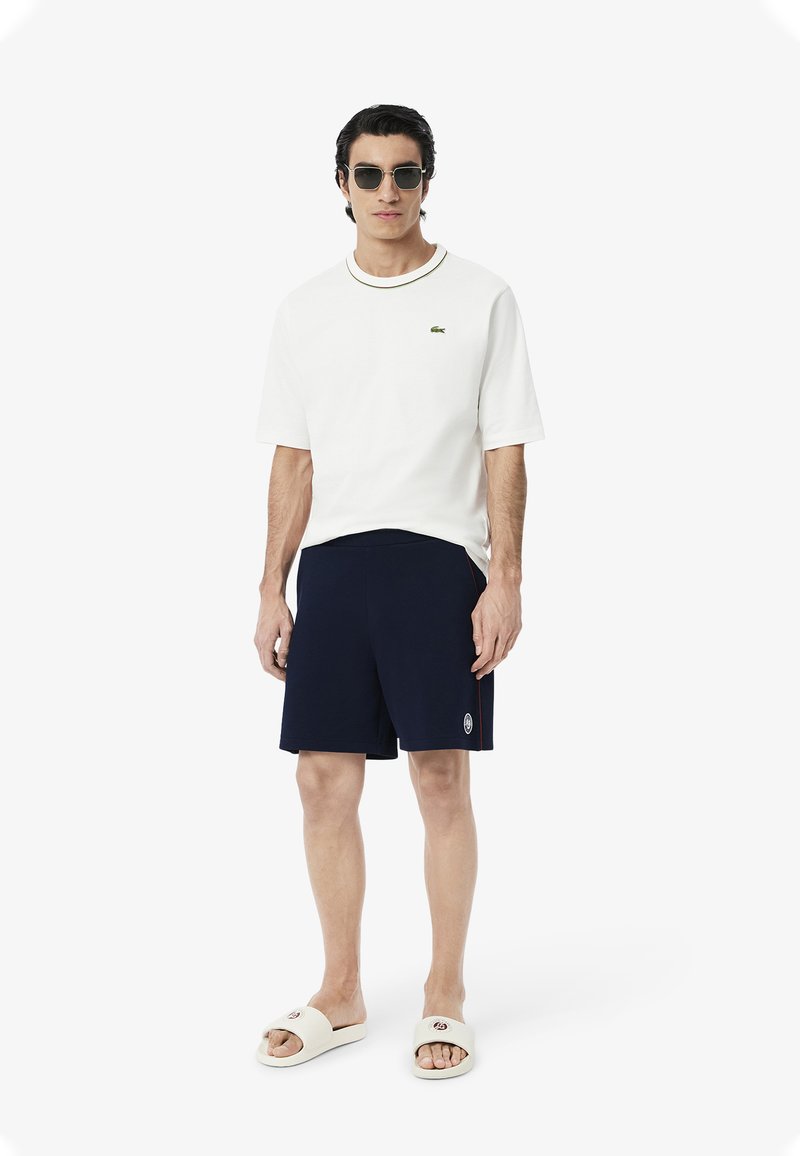 White short-sleeve t-shirt with a round neckline, navy shorts, and cream slide sandals. Reflective sunglasses complement the outfit.