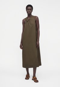 Femme portant une robe midi olive sans manches avec un détail nœud à l'épaule, associée à des sandales ouvertes marron.
