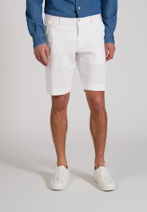 Man draagt witte knielange korte broek, witte sneakers en een blauw overhemd met lange mouwen, staand tegen een lichte achtergrond.
