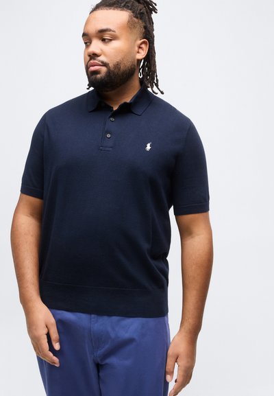 Άνδρας με ντραντλόκς που φοράει μπλε σκούρο πουκάμισο πόλο με κοντά μανίκια και μπλε παντελόνι, κοιτάζοντας προς τα αριστερά του, μπροστά σε ανοιχτό φόντο.