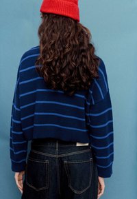 Maglione a righe blu navy con design corto. Presenta larghe strisce blu e polsini a costine. Abbinato a jeans in denim scuro.