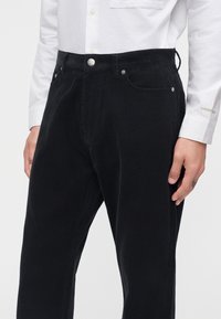 Homme portant un pantalon en velours côtelé noir et une chemise blanche à manches longues avec une poche devant, debout, mains détendues le long du corps.
