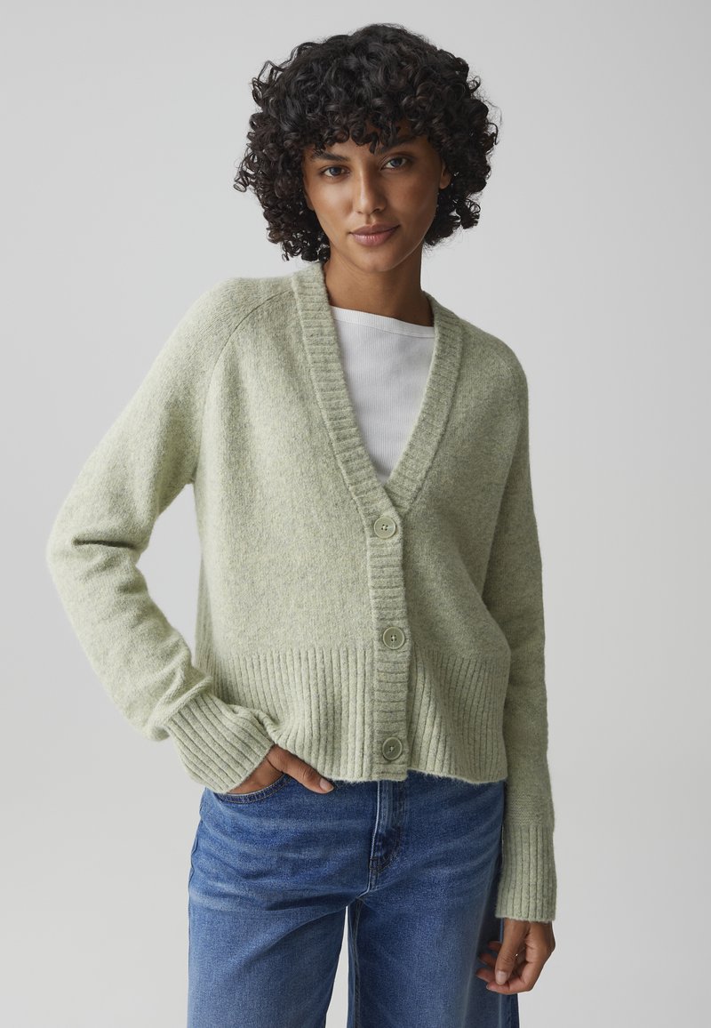 Femme aux cheveux bouclés portant un cardigan boutonné vert clair sur un haut blanc et un jean bleu, debout avec une main dans la poche.
