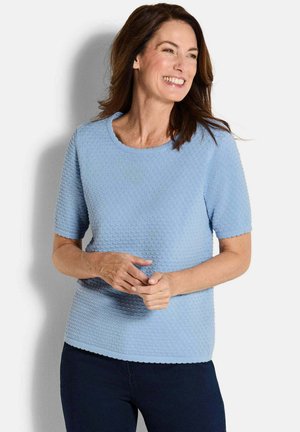 Donna che sorride e guarda di lato, indossando una maglietta azzurra chiara a maniche corte con texture e pantaloni scuri, in piedi davanti a uno sfondo semplice.