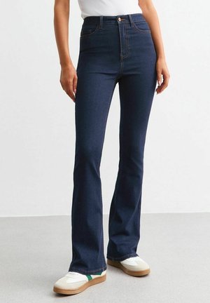 Personne portant un jean évasé taille haute bleu foncé et des baskets blanches avec une bande verte, debout sur un sol gris contre un mur blanc.