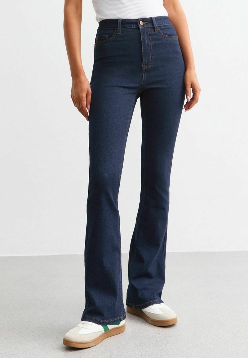 Personne portant un jean évasé taille haute bleu foncé et des baskets blanches avec une bande verte, debout sur un sol gris contre un mur blanc.
