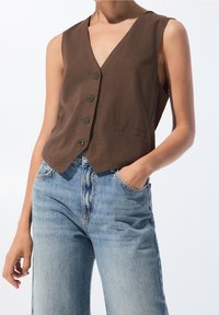 Gilet marron sans manches boutonné, porté avec un jean bleu clair taille haute, main droite du modèle dans la poche, bras gauche détendu le long du corps.
